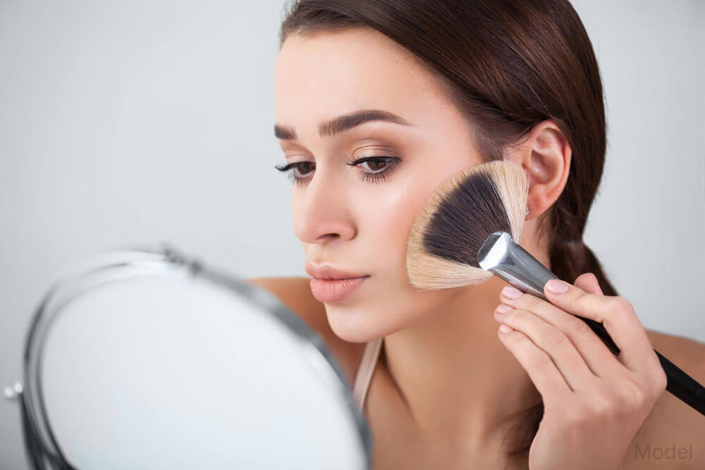 Female model brushing makeup on her face while looking into a mirror